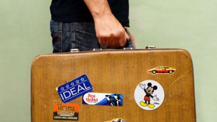 A leather suitcase carried by a man has stickers for travel destinations