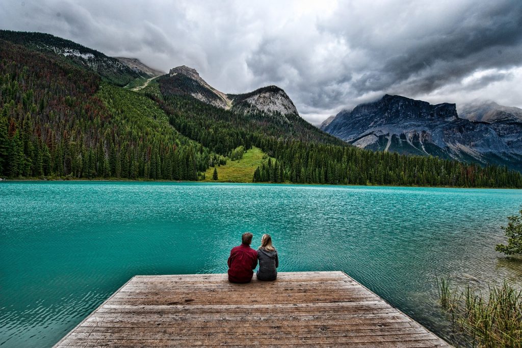 A man and a woman sit on a dock in front of a serene lake. This photo is for the guide to travel insurance for trips in Canada for Visitors to Canada.